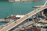 A view of a vibrant port area features a large bridge extending over a body of turquoise water. Cars drive along the bridge while parked vehicles are visible below. Numerous shipping containers are lined up in an organized manner alongside various buildings. A crane is positioned on the dock, and there's a sense of activity and movement in the harbor setting.