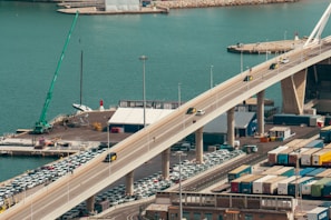 A view of a vibrant port area features a large bridge extending over a body of turquoise water. Cars drive along the bridge while parked vehicles are visible below. Numerous shipping containers are lined up in an organized manner alongside various buildings. A crane is positioned on the dock, and there's a sense of activity and movement in the harbor setting.