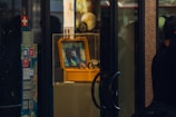 A display window features a yellow protective case, which is open to show an electronic device with a screen showing colorful patterns or maps. The case is placed on a pedestal inside a glass display box. There are various stickers with social media logos and other signs on the side wall. Reflections of people and objects outside the display can be seen on the glass surfaces.