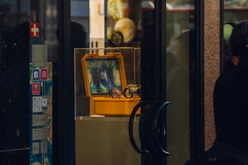 A display window features a yellow protective case, which is open to show an electronic device with a screen showing colorful patterns or maps. The case is placed on a pedestal inside a glass display box. There are various stickers with social media logos and other signs on the side wall. Reflections of people and objects outside the display can be seen on the glass surfaces.