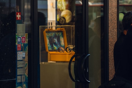 A display window features a yellow protective case, which is open to show an electronic device with a screen showing colorful patterns or maps. The case is placed on a pedestal inside a glass display box. There are various stickers with social media logos and other signs on the side wall. Reflections of people and objects outside the display can be seen on the glass surfaces.
