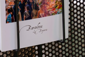 A postcard displays an image of a busy market scene with people walking between colorful stalls laden with a variety of fruits and vegetables. The text on the postcard reads 'Barcelona La Boqueria' in elegant script.