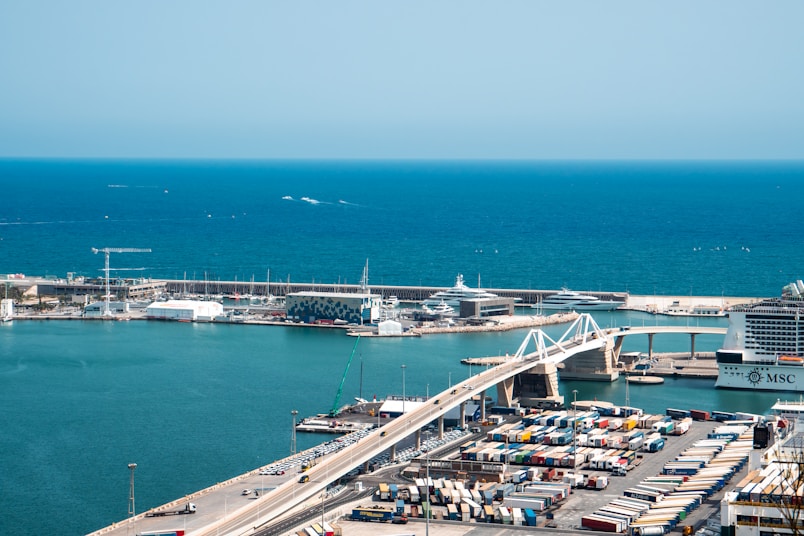 A large port area with numerous shipping containers arranged in rows is visible. There is a prominent cruise ship with the letters 'MSC' docked by a modern bridge. The background features a deep blue sea and yachts moored along the harbor.