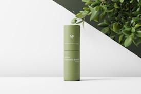 A green cylindrical cosmetic bottle with text on its surface, positioned on a minimalist white and dark green background. There are some leafy branches in the top right corner, adding a natural element to the composition.
