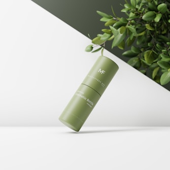 A green cylindrical cosmetic bottle is floating in a minimalist, well-lit setting. The background features a white surface with dark green foliage from a plant entering the frame from the upper right corner.