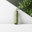 A cylindrical green cosmetic bottle standing on a diagonal surface with lush green leaves in the background. The bottle features minimalistic branding and appears to be part of an elegant product display.