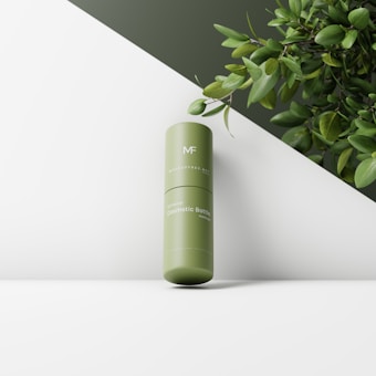 A cylindrical green cosmetic bottle standing on a diagonal surface with lush green leaves in the background. The bottle features minimalistic branding and appears to be part of an elegant product display.