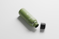 A reusable green water bottle lies on its side with a black screw-on cap beside it. The bottle features a subtle design with a silver handle attached to its neck.