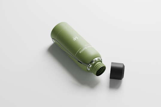 A reusable green water bottle lies on its side with a black screw-on cap beside it. The bottle features a subtle design with a silver handle attached to its neck.
