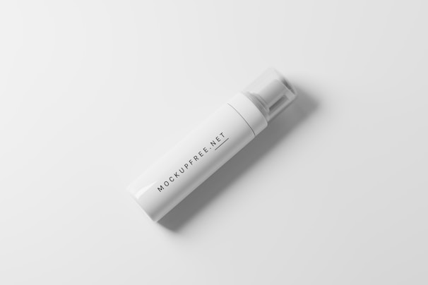 A white, cylindrical foam dispenser bottle is placed against a smooth, light gray background. The bottle features simple black text that reads 'MOCKUPFREE.NET' along its body. It has a clear plastic cap.