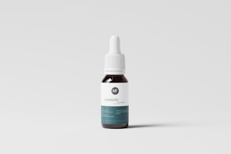 A small dropper bottle with a dark body and a white label featuring minimal text and a logo. The bottle has a white screw-on dropper cap, and it is positioned against a plain, light-colored background.