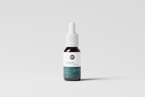 A small dropper bottle with a dark body and a white label featuring minimal text and a logo. The bottle has a white screw-on dropper cap, and it is positioned against a plain, light-colored background.