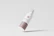 A sleek, minimalist cosmetic bottle tilted against a light gray background. The bottle is primarily white with a beige label at the bottom and has a spray nozzle at the top. The design is modern and clean, highlighting the branding in a simple, elegant manner.