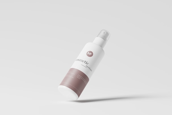 A sleek, minimalist cosmetic bottle tilted against a light gray background. The bottle is primarily white with a beige label at the bottom and has a spray nozzle at the top. The design is modern and clean, highlighting the branding in a simple, elegant manner.