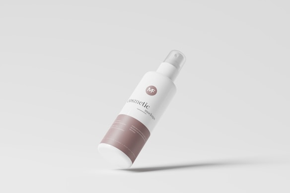 A sleek, minimalist cosmetic bottle tilted against a light gray background. The bottle is primarily white with a beige label at the bottom and has a spray nozzle at the top. The design is modern and clean, highlighting the branding in a simple, elegant manner.