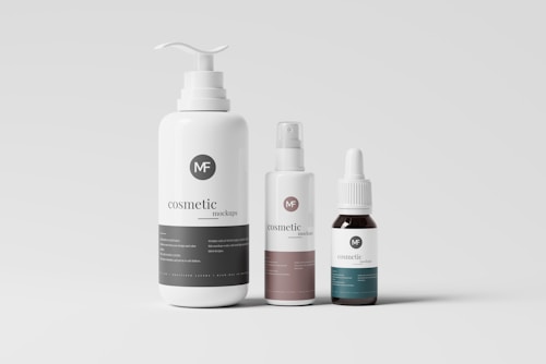 A minimalistic display of three cosmetic bottles with different shapes and sizes set against a plain background. The largest bottle has a pump dispenser, the middle one features a spray nozzle, and the smallest has a dropper cap. Each bottle has a simple, elegant label with the text 'cosmetic mockups' and a logo.