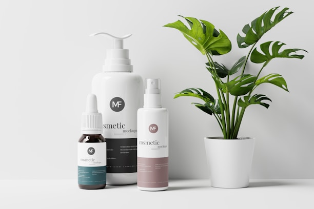 A minimalist arrangement of three cosmetic products next to a potted plant with green leaves. The cosmetics include a small dropper bottle, a pump bottle, and a spray bottle, all featuring a sleek, modern design with labels that read 'cosmetic mockups'. The potted plant adds a natural element to the scene, creating a balance between the products and the decor.