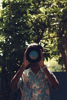 A person holds a vinyl record in front of their face, obscuring their identity. They are wearing a Hawaiian-style shirt with a tropical leaf pattern and a white watch on their wrist. The background is a lush and leafy outdoor setting, with sunlight filtering through the greenery.