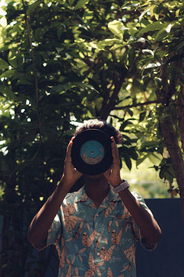 A person holds a vinyl record in front of their face, obscuring their identity. They are wearing a Hawaiian-style shirt with a tropical leaf pattern and a white watch on their wrist. The background is a lush and leafy outdoor setting, with sunlight filtering through the greenery.