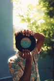 A person wearing a tropical print shirt holds up a vinyl record in front of their face. The background is filled with lush green foliage, creating a serene and natural setting. Sunlight filters through the leaves, casting a soft glow around the scene.