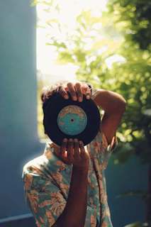 A person wearing a tropical print shirt holds up a vinyl record in front of their face. The background is filled with lush green foliage, creating a serene and natural setting. Sunlight filters through the leaves, casting a soft glow around the scene.