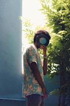 A person wearing a tropical-patterned shirt holds a vinyl record in front of their face while standing next to lush green foliage. Sunlight filters through the leaves, casting soft shadows on a pastel-colored wall in the background.