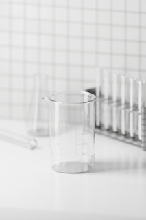 A clear glass beaker on a white surface with a grid-patterned wall in the background. The beaker is marked with measurements and is surrounded by glass test tubes and a test tube rack, creating a minimalistic and clean laboratory setting.