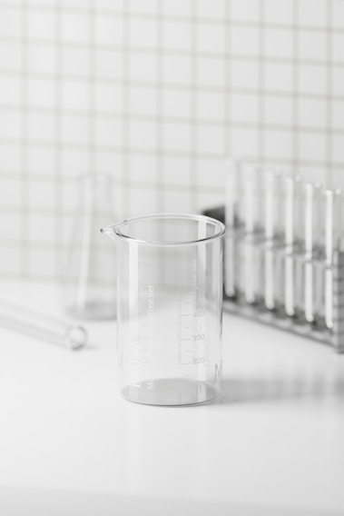 A clear glass beaker on a white surface with a grid-patterned wall in the background. The beaker is marked with measurements and is surrounded by glass test tubes and a test tube rack, creating a minimalistic and clean laboratory setting.