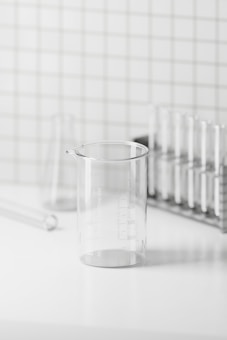 A clear glass beaker on a white surface with a grid-patterned wall in the background. The beaker is marked with measurements and is surrounded by glass test tubes and a test tube rack, creating a minimalistic and clean laboratory setting.