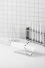 A clear glass beaker is lying on its side with visible measurement markings on a white surface. In the background, there is a metal rack holding several test tubes. The setting appears to be minimalistic, with a grid-patterned wall.