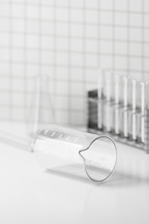 A clear glass beaker is lying on its side with visible measurement markings on a white surface. In the background, there is a metal rack holding several test tubes. The setting appears to be minimalistic, with a grid-patterned wall.