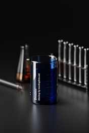 A dark-themed laboratory scene featuring a variety of glassware. Prominently in the foreground is a blue beaker with measurement markings and the text 'Beaker' printed on it. Behind, there are test tubes organized in a rack and a conical flask with an amber-colored liquid. The setting conveys a scientific and industrial vibe.
