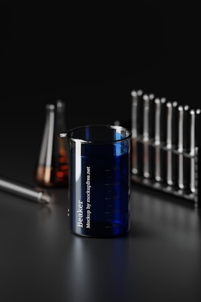 A dark-themed laboratory scene featuring a variety of glassware. Prominently in the foreground is a blue beaker with measurement markings and the text 'Beaker' printed on it. Behind, there are test tubes organized in a rack and a conical flask with an amber-colored liquid. The setting conveys a scientific and industrial vibe.