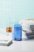 A transparent glass beaker filled with blue liquid is placed on a white surface near a stack of books. In the background, there is a blurred view of a conical flask containing an amber-colored liquid, and another scattered piece of laboratory glassware. The background wall has a light blue tile pattern.