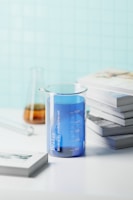 A transparent glass beaker filled with blue liquid is placed on a white surface near a stack of books. In the background, there is a blurred view of a conical flask containing an amber-colored liquid, and another scattered piece of laboratory glassware. The background wall has a light blue tile pattern.