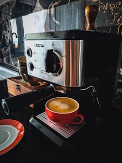 A Nova espresso machine sits on a kitchen counter with a freshly brewed coffee in a vibrant red cup resting on its drip tray. The machine is sleek with a metallic finish, featuring a clear control panel. A decorative wooden box and a white saucer with a red rim accompany the setup, adding a touch of elegance. The backdrop is a sophisticated marble wall, enhancing the overall luxurious feel.