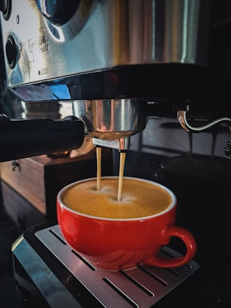 A red cup is positioned under a coffee machine, with coffee being dispensed into it. The machine is metallic and has a glossy surface with several buttons and an espresso filter. The background features a wooden box and the reflection of the surroundings can be seen on the machine.