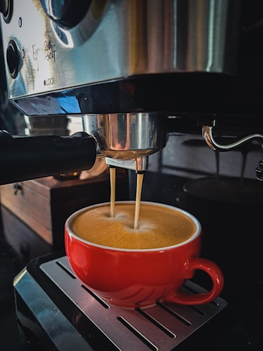 A red cup is positioned under a coffee machine, with coffee being dispensed into it. The machine is metallic and has a glossy surface with several buttons and an espresso filter. The background features a wooden box and the reflection of the surroundings can be seen on the machine.