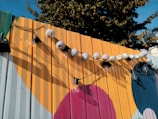 A wooden fence painted with large, colorful geometric shapes serves as a backdrop for a string of round paper lanterns and exposed lightbulbs. The warm colors of the fence contrast with the blue sky and greenery visible in the background.