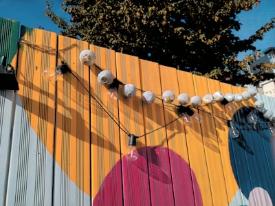 A wooden fence painted with large, colorful geometric shapes serves as a backdrop for a string of round paper lanterns and exposed lightbulbs. The warm colors of the fence contrast with the blue sky and greenery visible in the background.