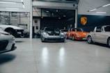 A collection of vintage and modern sports cars are parked inside a spacious, well-lit garage with polished concrete floors. The vehicles include a classic orange car and a sleek, modern black car at the center. The environment appears clean and organized, with a focus on high-performance automobiles. A large Porsche emblem is displayed on the wall, indicating the brand of the cars.