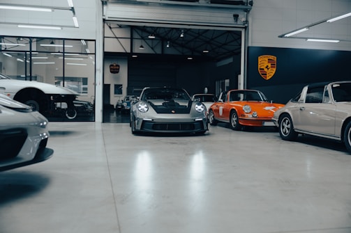 A collection of vintage and modern sports cars are parked inside a spacious, well-lit garage with polished concrete floors. The vehicles include a classic orange car and a sleek, modern black car at the center. The environment appears clean and organized, with a focus on high-performance automobiles. A large Porsche emblem is displayed on the wall, indicating the brand of the cars.