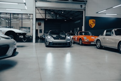 A collection of vintage and modern sports cars are parked inside a spacious, well-lit garage with polished concrete floors. The vehicles include a classic orange car and a sleek, modern black car at the center. The environment appears clean and organized, with a focus on high-performance automobiles. A large Porsche emblem is displayed on the wall, indicating the brand of the cars.