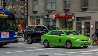 A busy city street scene featuring a green taxi, a black SUV, and a blue city bus. Several storefronts are visible, including a Starbucks, a jewelry store, and a market with a red sign. A cyclist is also nearby, and urban buildings form the backdrop.