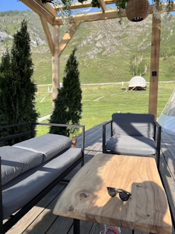 A wooden outdoor seating area with a table and two cushioned chairs situated under a pergola. The surrounding landscape features green grassy hills and distant mountains. A pair of sunglasses rests on the table. Potted evergreens are placed near the seating area, and a rattan-style hanging basket is visible overhead.