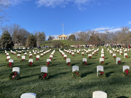 An expansive cemetery with rows of white tombstones, each adorned with red wreaths, stretches across a green field. In the distance, a neoclassical building with columns stands on a small hill, with a flagpole bearing an American flag in front of it. A crowd of people gathers near the building and among the trees in the background under a clear blue sky.