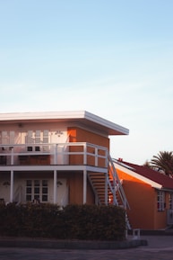 A modern two-story house with a flat roof and vibrant orange walls. The upper level features a balcony with white railings, and there is an external staircase leading down to the ground level. Below the house, there is a hedge and a paved area. The sky is clear, and there are some palm trees in the background.