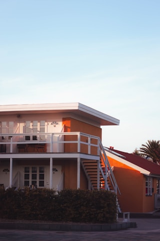 A modern two-story house with a flat roof and vibrant orange walls. The upper level features a balcony with white railings, and there is an external staircase leading down to the ground level. Below the house, there is a hedge and a paved area. The sky is clear, and there are some palm trees in the background.