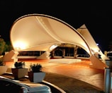A modern architectural structure featuring a large, illuminated canopy with a smooth, curved design, set against a dark night sky. The structure is supported by robust, angular beams, and is surrounded by potted plants and a parked vehicle with reflective surfaces. The ground consists of terracotta tiles, adding warmth to the scene.