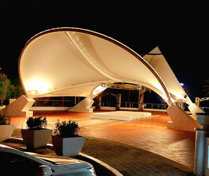 A modern architectural structure featuring a large, illuminated canopy with a smooth, curved design, set against a dark night sky. The structure is supported by robust, angular beams, and is surrounded by potted plants and a parked vehicle with reflective surfaces. The ground consists of terracotta tiles, adding warmth to the scene.